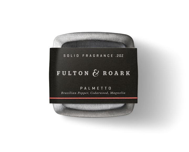 A sleek, metallic black container with rounded edges, labeled "Fulton & Roark," holds the Palmetto .2oz solid fragrance. This fragrance features notes of Brazilian Pepper, Cedarwood, and Magnolia.