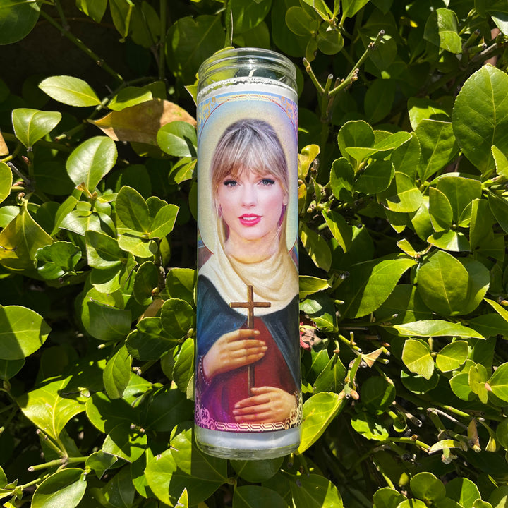 The Taylor Swift Candle from BOBBYK BOUTIQUE is a tall devotional featuring an illustration of a blonde woman with bangs, wearing a white head covering, and holding a cross and book. The unscented candle is set against a lush green leafy background.