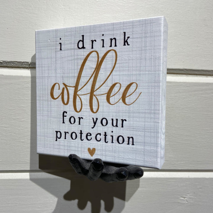 In Rock Rapids, IA, a small sign elegantly displays the phrase "I drink coffee for your protection" in stylish fonts, accompanied by a tiny heart symbol at the bottom. This handcrafted design from SincereSurroundings is named "Coffee Protection" and is held gracefully by a black decorative hand-shaped shelf display.