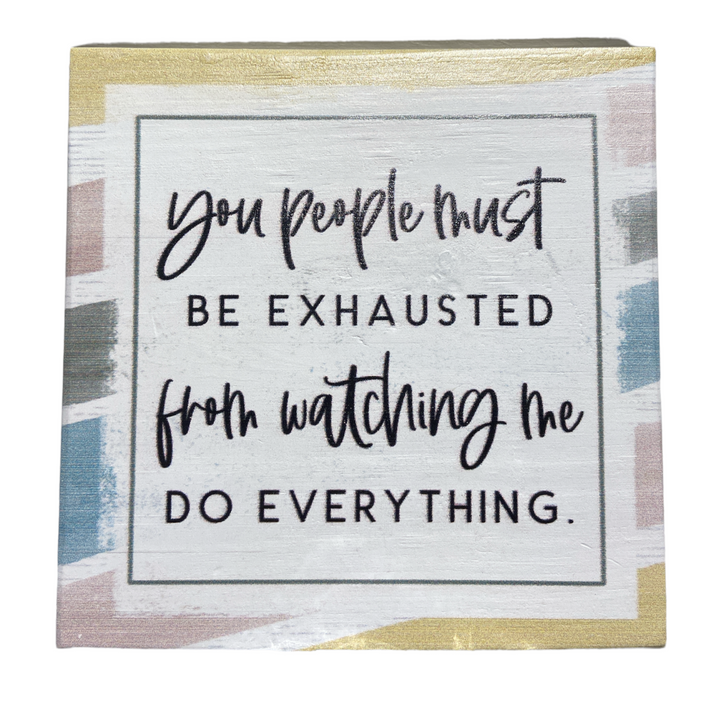 A handcrafted decorative sign from SincereSurroundings, titled "You People," features the phrase "You people must be exhausted from watching me do everything" on a pastel geometric pattern. It's an ideal addition to your display shelf, evoking the charm of a Rock Rapids, IA find.