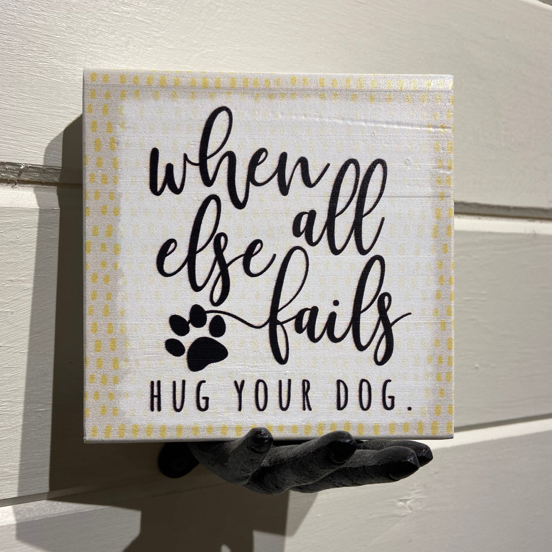 A unique decorative piece by SincereSurroundings, the "Hug Your Dog" sign features the heartfelt message, "When all else fails hug your dog," alongside a charming paw print illustration. Stylishly presented on a black sculpted hand with a white wooden backdrop, it adds an ideal touch to any shelf display in Rock Rapids homes.