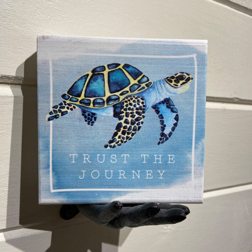 The "Trust The Journey" artwork by SincereSurroundings is a handcrafted canvas showcasing a blue and yellow sea turtle alongside the words "TRUST THE JOURNEY." Designed for effortless display, it sits gracefully on a small hand sculpture set against a light-colored wooden backdrop.