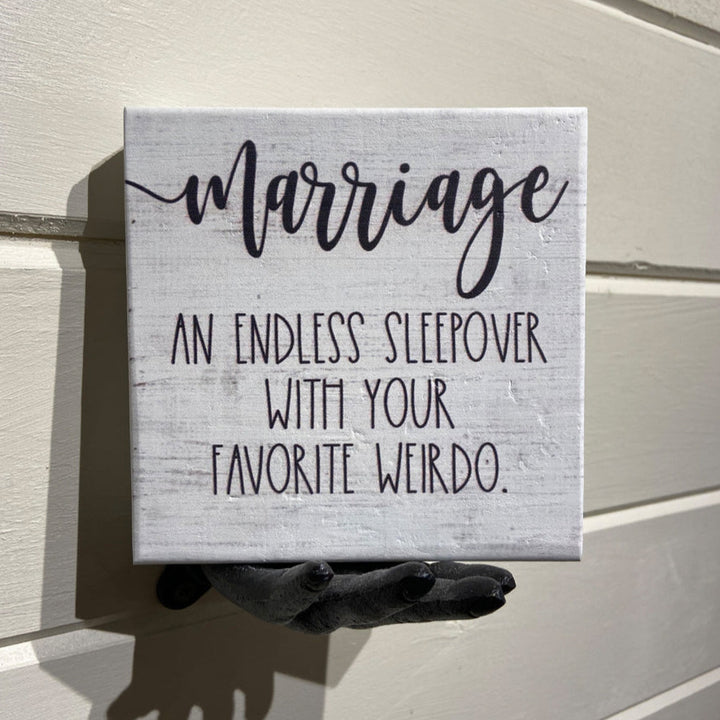 A small sign from SincereSurroundings, named "Marriage Endless," featuring the text "An endless sleepover with your favorite weirdo," is Easy Display mounted on a white wall. A black handcrafted hand sculpture appears to hold the sign from below.