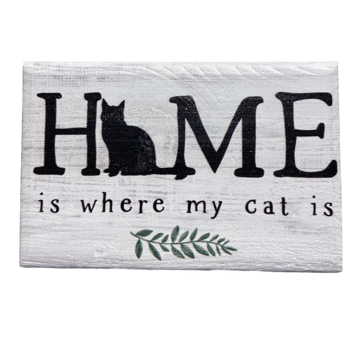 The "Home Is Where Cat" sign by SincereSurroundings is a handcrafted wooden piece featuring the phrase "HOME is where my cat is" in black text. The letter "O" in "HOME" includes a silhouette of a sitting cat, accompanied by a small green leaf graphic below. This charming display boasts a rustic white-washed finish and is made in Rock Rapids, IA.