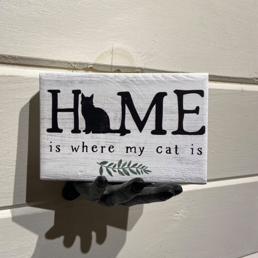 The SincereSurroundings "Home Is Where Cat" decor piece features a small wooden sign held by a black sculpted hand. The sign showcases the phrase "HOME is where my cat is," with a silhouette of a sitting cat incorporated into the word "HOME" and a small green twig illustration below the text. This handcrafted item adds charm to any display.