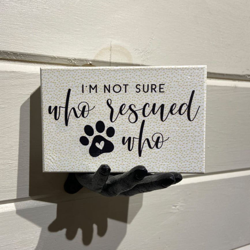 The "Who Rescued Who" plaque by SincereSurroundings features a heartfelt message in black script font, reading "I'm not sure who rescued who." Below the text is a charming paw print with a heart at its center. This handcrafted piece is easy to display, resting on a black hand-shaped wall hook against a white textured wall. Made in Rock Rapids.