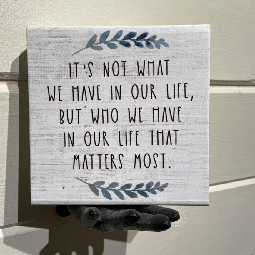 A small handcrafted plaque from SincereSurroundings, named "Who We Have," featuring the quote "It's not what we have in our life, but who we have in our life that matters most," with leaf illustrations, is artfully positioned on a shelf display. This unique piece adds charm to any Rock Rapids home.