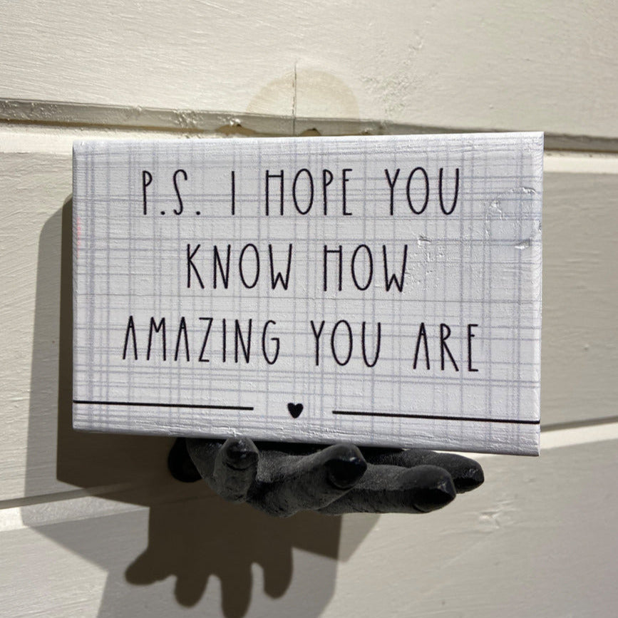 A small rectangular sign from SincereSurroundings called "P.S. Amazing" is mounted on a wall with a light gray plaid background and black text reading, "P.S. I hope you know how amazing you are," followed by a tiny heart symbol. Ideal for your Rock Rapids shelf display, the sign hangs from a handcrafted black hand-shaped hook.