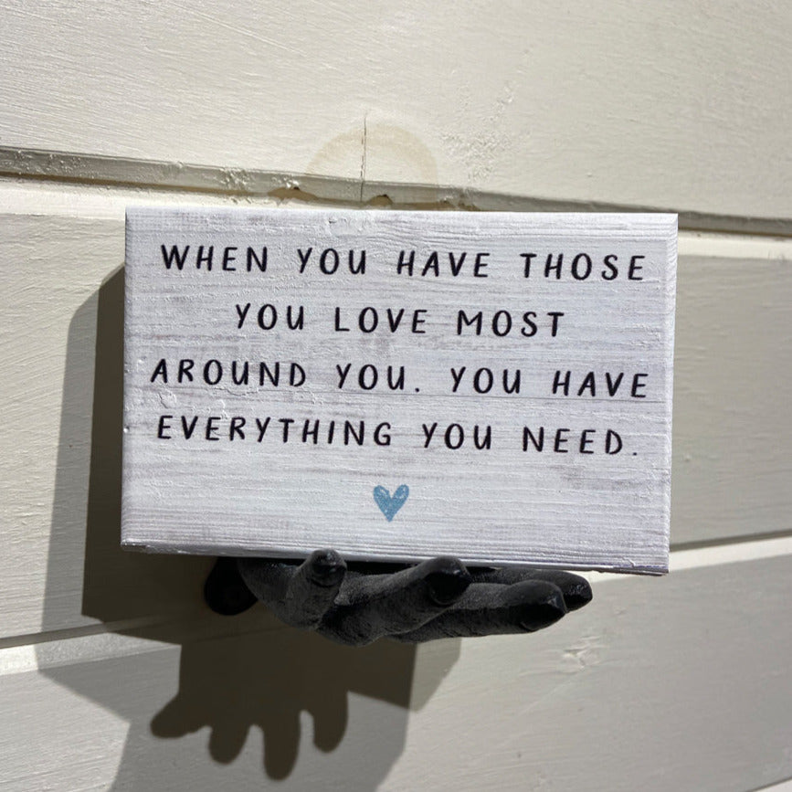 A small, handcrafted sign from SincereSurroundings called "Those You Love," featuring the message "When you have those you love most around you, you have everything you need," is written in black text on a white background. The sign is mounted on a wall in Rock Rapids and supported by a gray hand sculpture with a small blue heart symbol below the text.