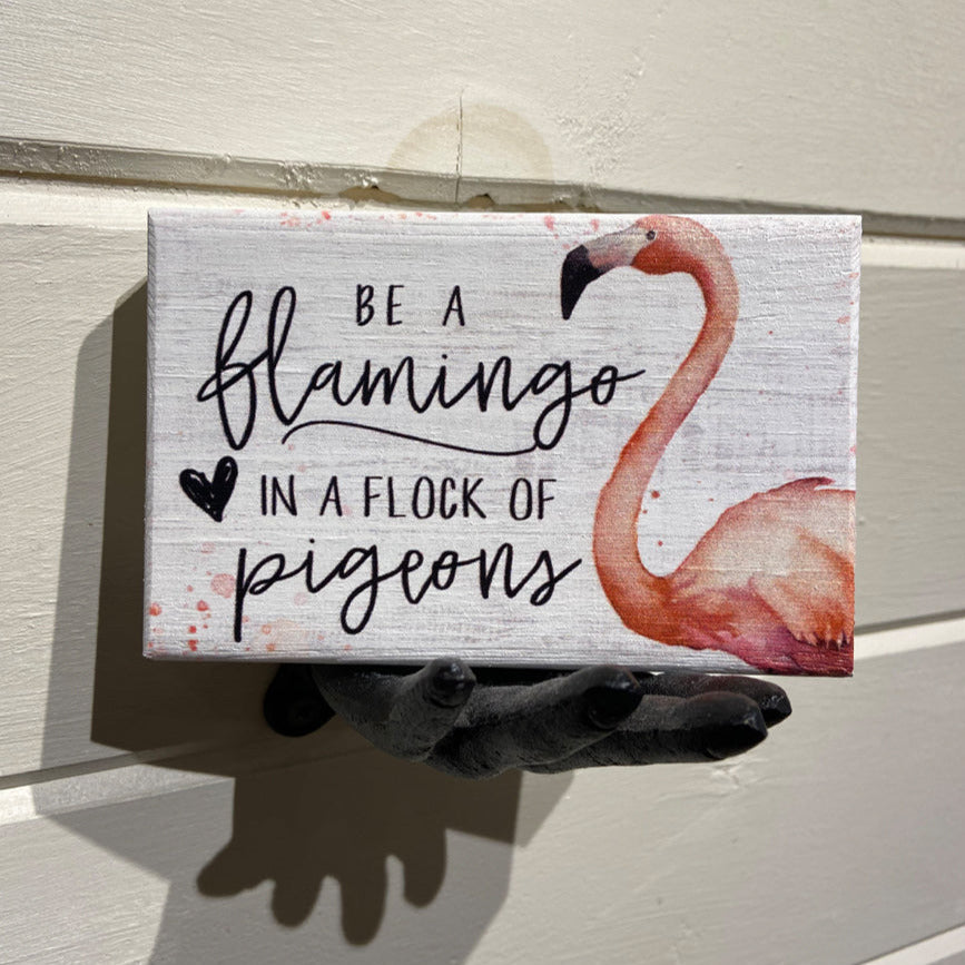 A crafted shelf display features the "Be A Flamingo" sign by SincereSurroundings, showcasing a painted pink flamingo with the phrase "Be a flamingo in a flock of pigeons" and adorned with a small black heart, elegantly held by a hand sculpture from Rock Rapids.