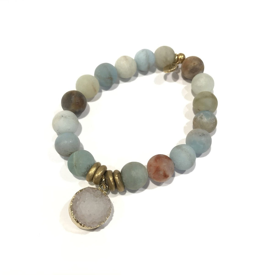 The Lenny & Eva 10mm Gemstone Amazonite beaded bracelet features round, multicolored stones in shades of blue, brown, and gray. It includes several brass accents and a gold-framed druzy quartz charm hanging from the center. The bracelet is set against a white background and stretches to fit most wrists.