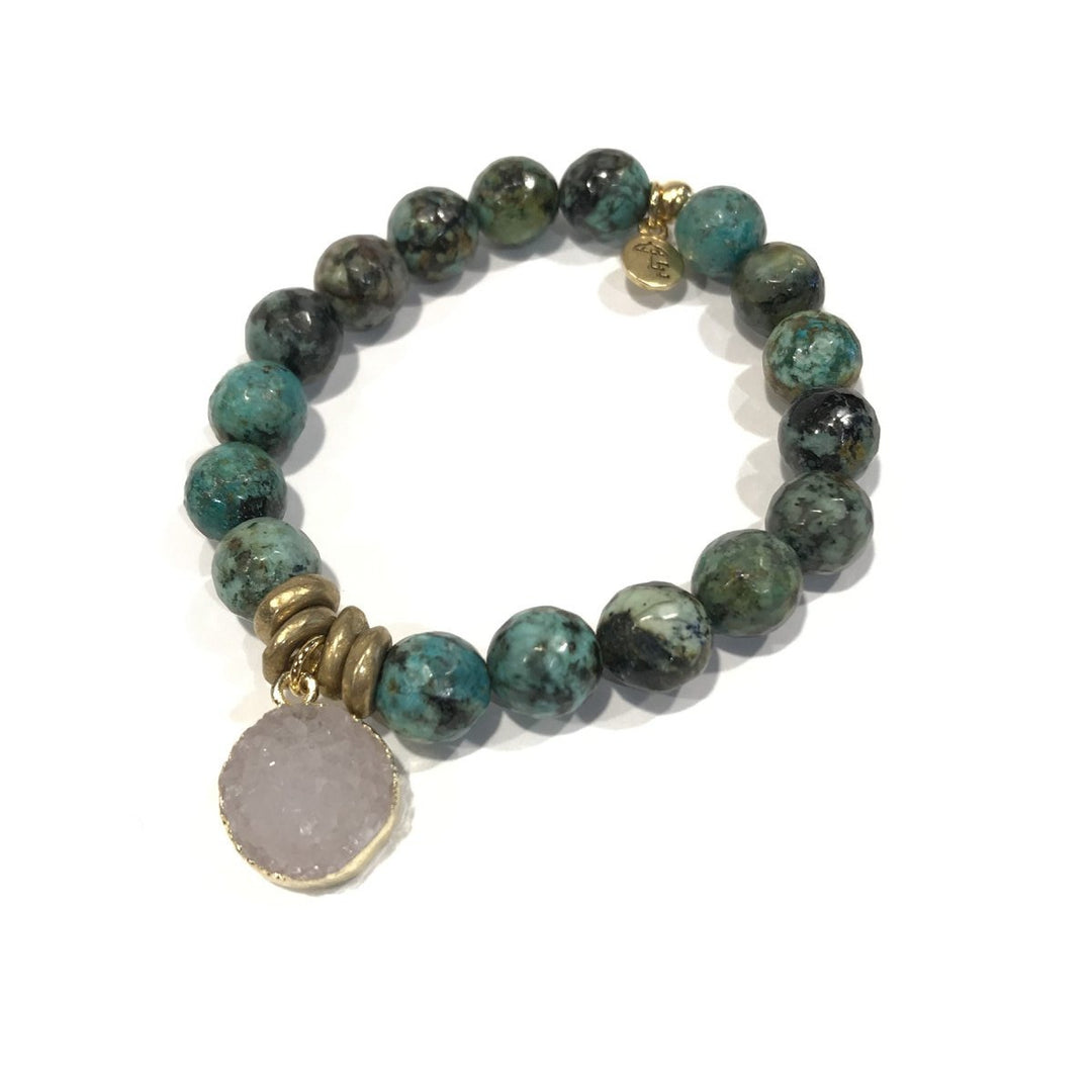 The Lenny & Eva beaded bracelet features 10mm African turquoise and green natural semi-precious gemstones, complemented by gold spacer beads, brass accents, and a circular crystal pendant framed with a gold rim. A small gold charm near the clasp adds an extra touch of elegance. The stretchable design ensures it fits most wrists, and it is showcased in a circular arrangement against a white background.