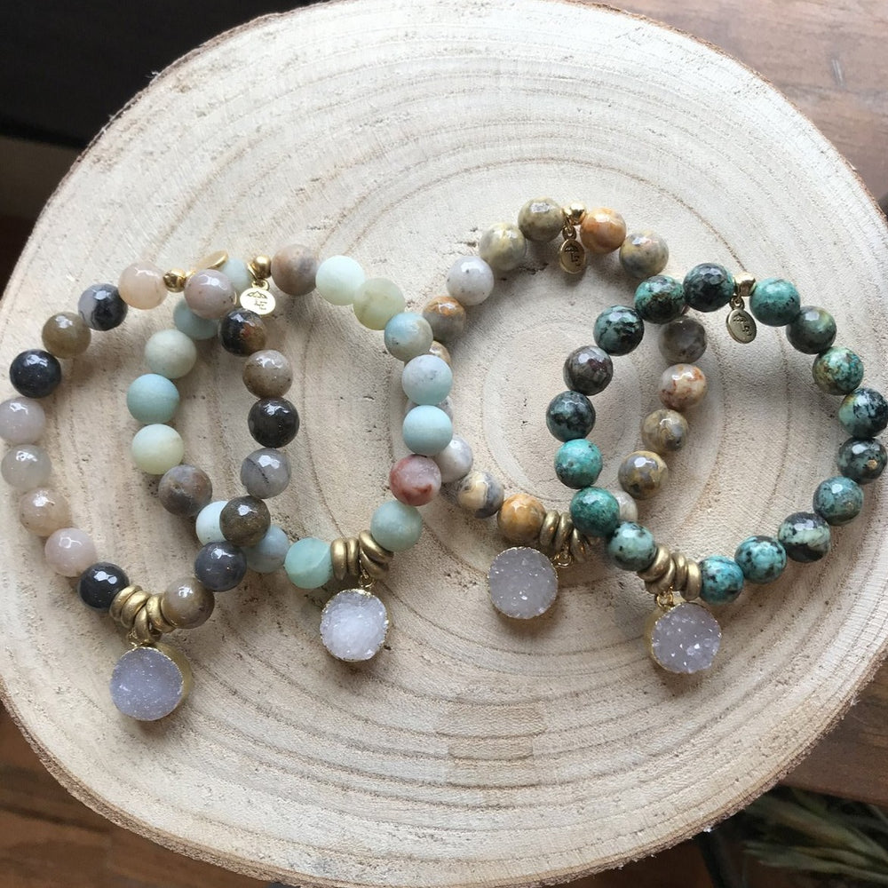 Four Lenny & Eva beaded bracelets featuring natural 10mm Amazonite gemstones and brass accents are displayed on a circular, light-colored wooden slab. Each bracelet has a charm with a circular stone pendant and stretches to fit most wrists. The background is a warm wooden surface.