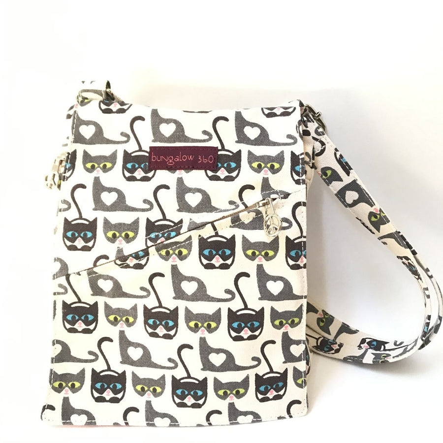 The Sm Messenger Cat from Bungalow 360 is a crossbody bag featuring unique designs with repeated prints of black and grey cats, each with green and yellow eyes wearing blue glasses, alongside white heart shapes. It includes a front zipper pocket and proudly carries the brand's name "Bungalow 360," known for being 100% vegan.