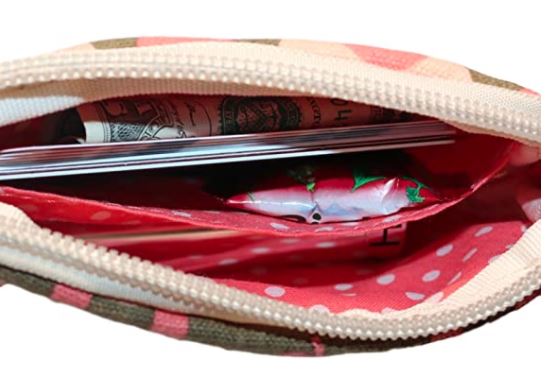 A close-up view of the interior of an open Bungalow 360 Tiny Wristlet Pig with a zipper. Inside the wristlet, there are neatly folded U.S. dollar bills, a few pens, and a small package with a colorful wrapper. The inner lining of the wristlet showcases unique designs featuring red with white polka dots.