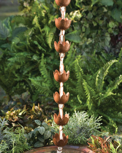 The Rain chain Lily by ANCIENTGRAFF features a vertical series of copper-plated, flower-shaped cups designed to elegantly guide rainwater downwards in a garden setting adorned with lush greenery. With adjustable lengths for customization, the chain is mounted above a water-catching basin using gutter clips.