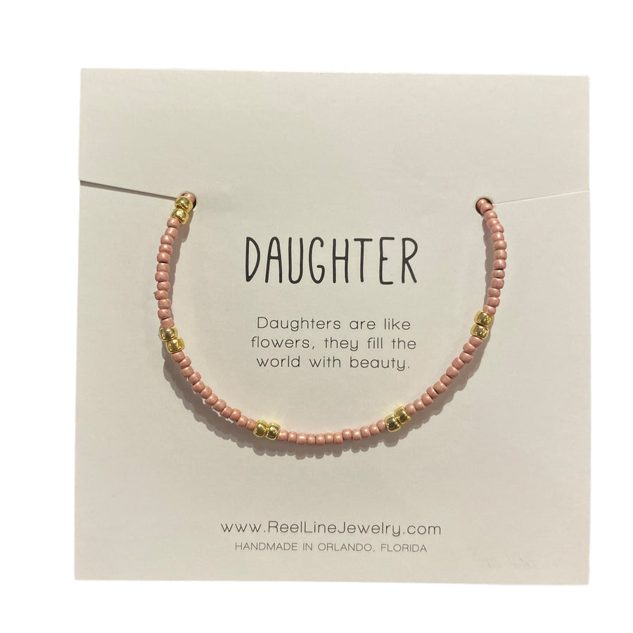 A fragile thin glass beaded bracelet in pink with gold accents, named "Daughter - Pink and Gold," is showcased on a card. The card states, "Daughters are like flowers, they fill the world with beauty," alongside the URL "www.ReelLineJewelry.com" and the note "Handcrafted in Florida" by Reel Line Jewelry. It features adjustable links for a perfect fit.