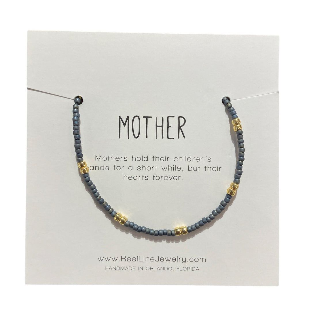 The "Mother - Denim and Gold" bracelet by Reel Line Jewelry features a handcrafted design with blue and gold glass beads. It is presented on a card that reads, "MOTHER - Mothers hold their children's hands for a short while, but their hearts forever." With adjustable links for the perfect fit, the bottom of the card includes website and location details.