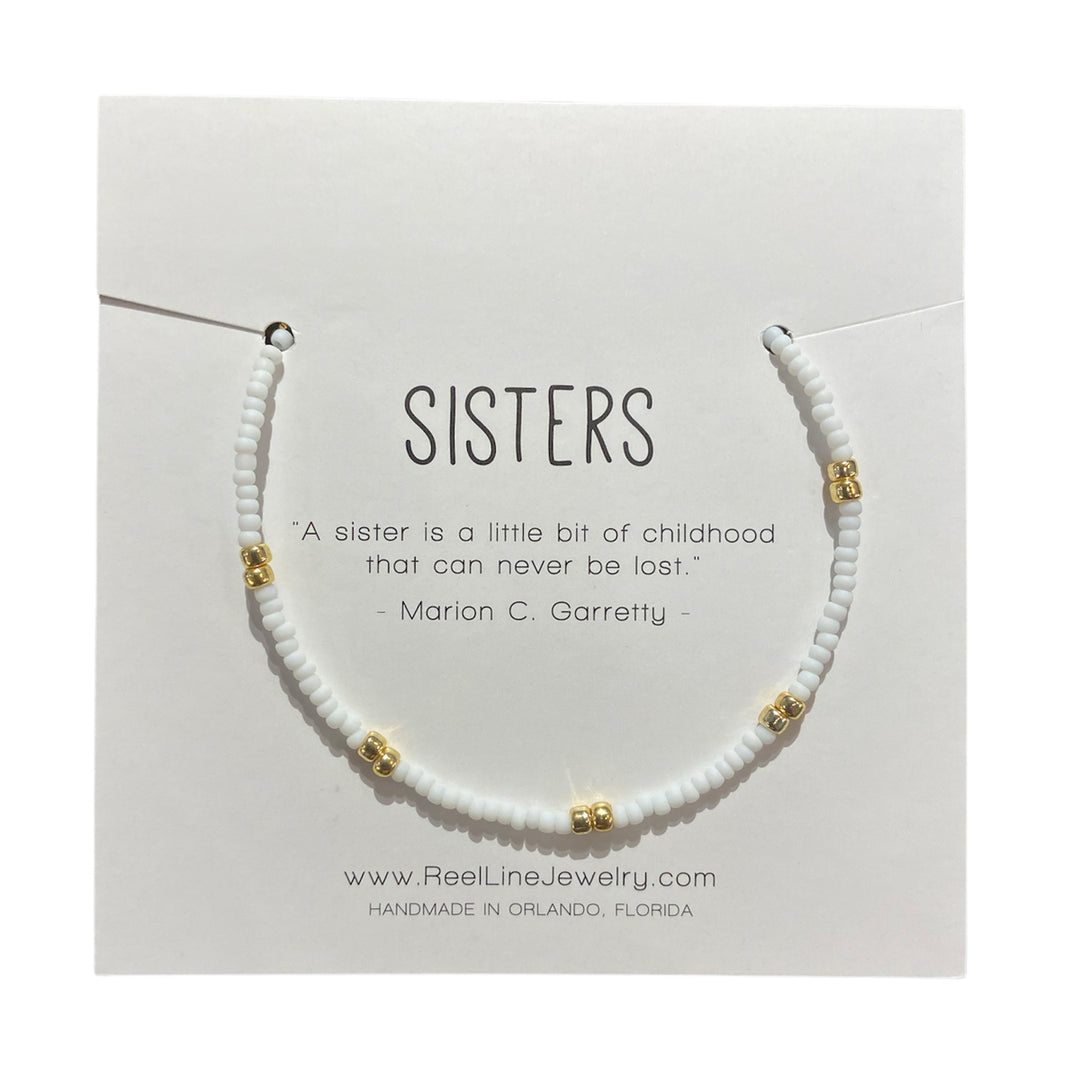 The "Sisters - Frost and Gold" bracelet by Reel Line Jewelry features delicate white and gold beads arranged on a thin glass band. It is elegantly displayed on a card inscribed with the quote: "SISTERS. 'A sister is a little bit of childhood that can never be lost.' - Marion C. Garretty." The card also notes, "Handcrafted in Orlando, Florida," along with the website www.ReelLineJewelry.com.