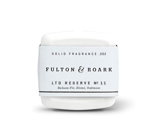 A white, rectangular tin of solid fragrance by Fulton & Roark, named "Ramble .2oz," boasts a captivating scent blend of Balsam Fir, Elemi, and Oakmoss. The tin features a minimalist design with black text on a light blue background.