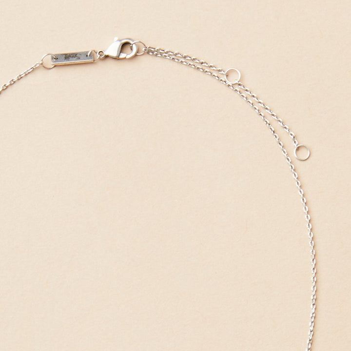 A piece from Scout Jewelry, the Refined Neck Crescent in silver, elegantly arranged in a partial circle on a plain beige background. This delicate necklace features a lobster clasp and a small rectangular tag near the clasp that highlights its exquisite craftsmanship.