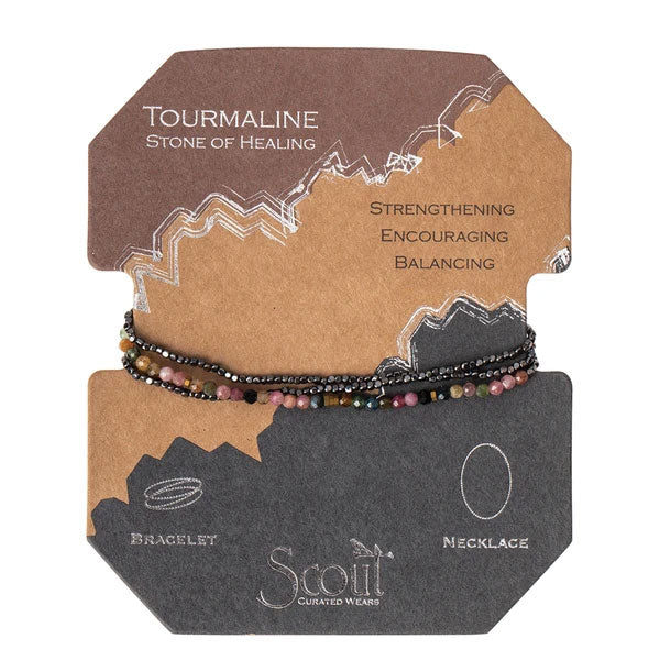 The Tourmaline Hematite Delicate Wrap by Scout Jewelry is a versatile set of tourmaline beads that can be worn as both a bracelet and a necklace. Strung on stretch cord, it is presented on an elegant brown and gray packaging card. The card reads "Tourmaline, Stone of Healing: Strengthening, Encouraging, Balancing" and features the brand name "Scout Curated Wears.