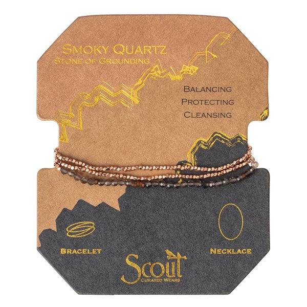 The Scout Jewelry Smoky Quartz/Rose Gold Delicate Wrap set includes a gemstone beaded bracelet and necklace. Packaging features balancing, protecting, cleansing benefits in brown/black cardboard with yellow accents and doodles.