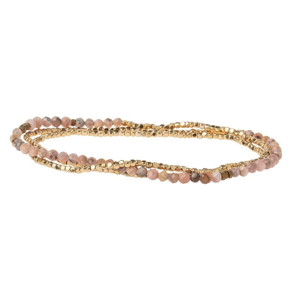 The Scout Jewelry Rhodochrosite/Gold Delicate Wrap is a chic bracelet showcasing alternating rows of small gold beads and rose-colored rhodochrosite gemstones, beautifully intertwined in a sleek and subtle design against a white backdrop.