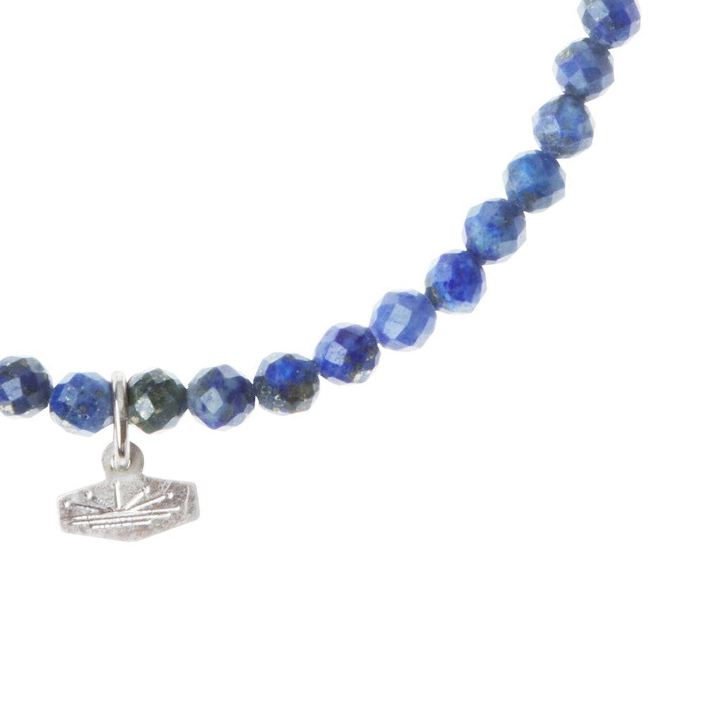 A close-up view of the Mini Bracelet Lapis/Silver by Scout Jewelry showcases round, faceted blue semi-precious stone beads. The bracelet features a small, silver, boat-shaped charm hanging at its center. The beads and charm are displayed against a white background.