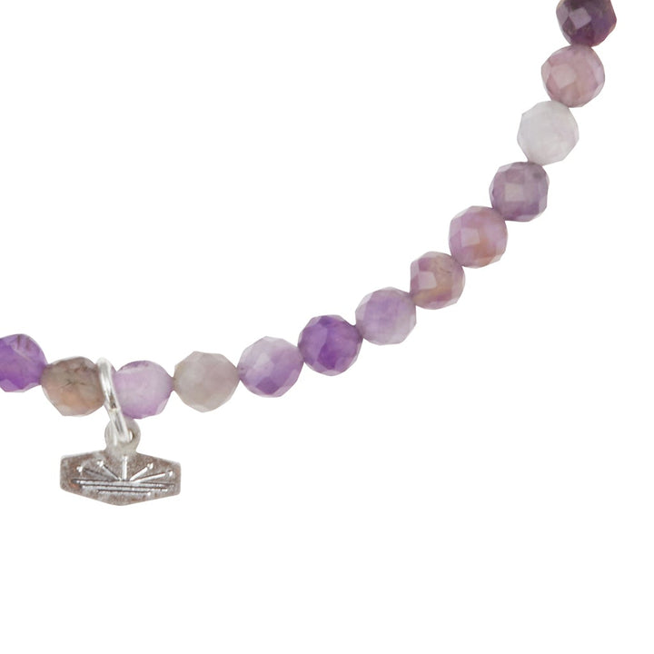 A close-up of the Mini Bracelet Amethyst/Silver by Scout Jewelry showcases faceted stone beads in shades of purple and lavender. A small silver charm with an engraved design adorns the piece, set against a white background.