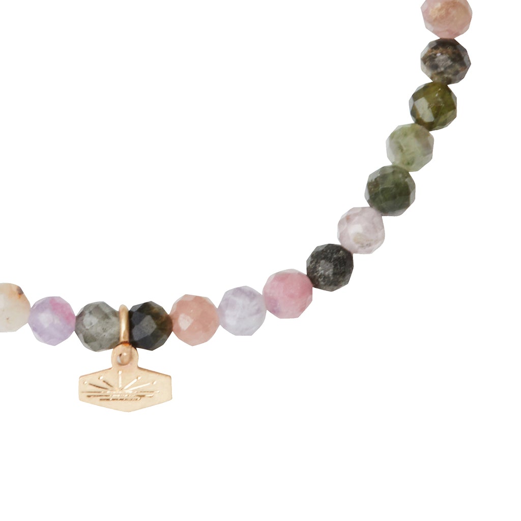 A detailed view of the Mini Bracelet Tourmaline/Gold by Scout Jewelry, featuring faceted, semi-precious gemstones in beautiful shades of pink, green, and purple. A delicate gold charm with an engraved design is elegantly suspended between two beads on the stretch cord. The background is white.