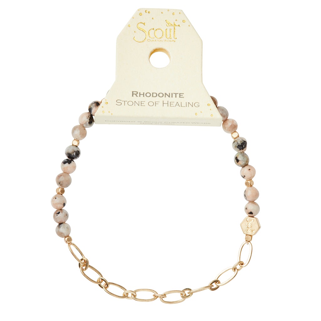 The Chain Bracelet Rhodonite/Gold by Scout Jewelry features round and oval pink and gray semi-precious gemstones on a stretch cord with a gold-tone paperclip chain accent. It is elegantly packaged as "Scout" with the Rhodonite Stone of Healing.