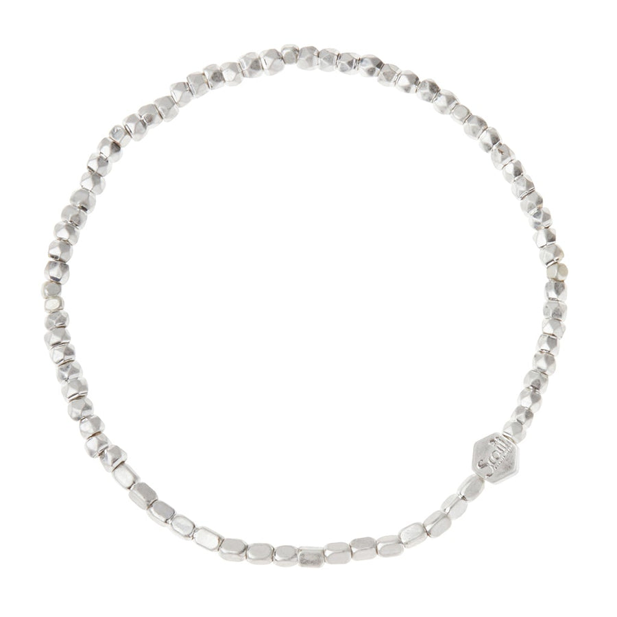 A close-up image of the Scout Jewelry Mini Metal Bracelet Silver, featuring small, irregularly shaped silver beads strung on a stretch cord. The circular bracelet includes a small hexagonal charm with text on it and showcases mixed metals for subtle accents against a plain white background.