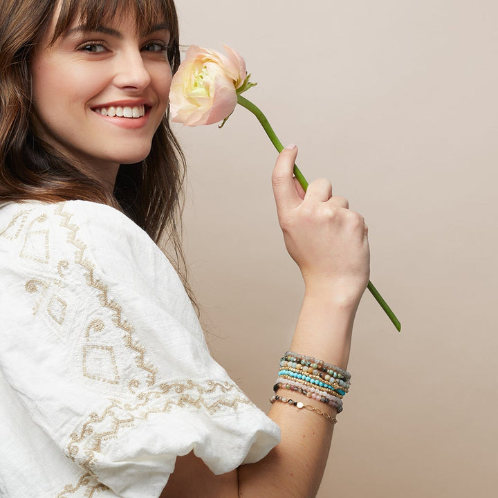 A woman with brown hair wears a white embroidered blouse and the Chain Bracelet Blue Sky Jasper/Silver by Scout Jewelry while holding a pink rose near her face, smiling against a beige background.