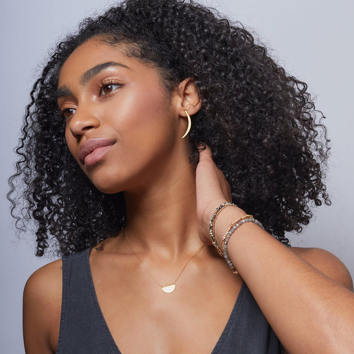 A person with curly black hair wears the Gibbous Slice Stud Silver from Scout Jewelry. Their necklace shines in matching 18k gold vermeil, and layered bracelets adorn their neck-touched left hand against a light gray background.