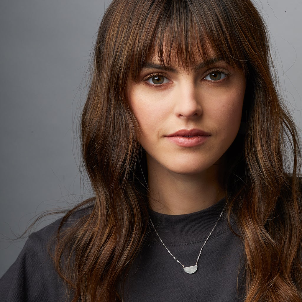 A young woman with long, wavy brown hair and bangs wears the Scout Jewelry Refined Neck Half Moon/Silver necklace, featuring a 925 silver half-circle pendant on an adjustable chain, paired with a dark top against a plain gray background.