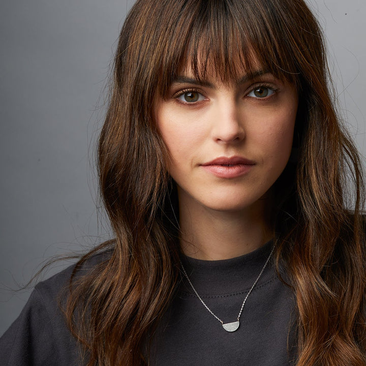 A young woman with long, wavy brown hair and bangs wears the Scout Jewelry Refined Neck Half Moon/Silver necklace, featuring a 925 silver half-circle pendant on an adjustable chain, paired with a dark top against a plain gray background.