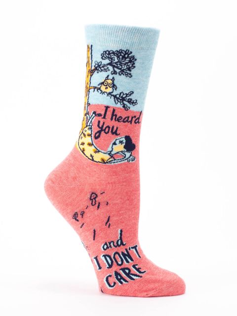 The Blue Q "I heard you and I don't care" socks showcase a cartoon design of a person lounging in a hammock tied to a tree branch, with an owl watching nearby. The socks feature a blue and pink color scheme and include the text "I heard you...and I don't care.