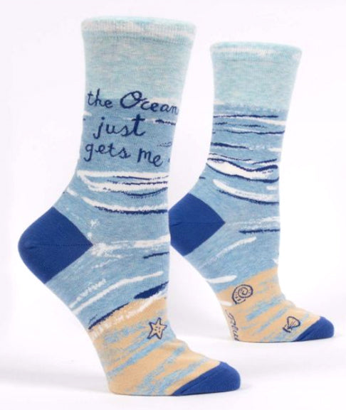 The "Ocean Gets Me Socks" by Blue Q are light blue with dark blue, white, and tan wave patterns. Crafted from cozy combed cotton, these women's shoe socks feature the text "the Ocean just gets me" near the top. The toes and heels are dark blue, while the soles display small sea-themed designs like a starfish and a shell.