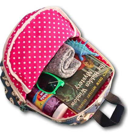 The Backpack Sea Otter by Bungalow 360 is an open cotton canvas bag with a polka dot lining, containing sunglasses, a rolled towel, a book titled "The Penderwicks," a toy dinosaur, a small jar labeled "Fruit Pouch," and a purple bottle. Each item adds charm to this unique design.