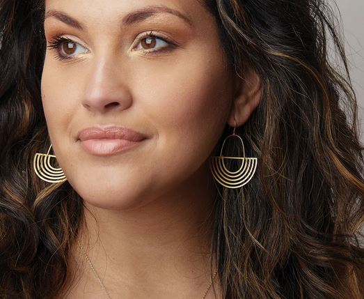 A woman with long, wavy hair is wearing large, geometric earrings called Solar Rays Silver from Scout Jewelry. She has a neutral expression and is looking off to the side. Her makeup is subtle, featuring warm-toned eyeshadow and a natural lip color.