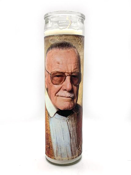 A tall, unscented decorative candle from BOBBYK BOUTIQUE, named the "Stan Lee Candle," features an image of an older man with white hair, wearing dark sunglasses, a light blue shirt, and a brown jacket. The background is a light tan color with subtle patterns, celebrating him as "Man the Legend.