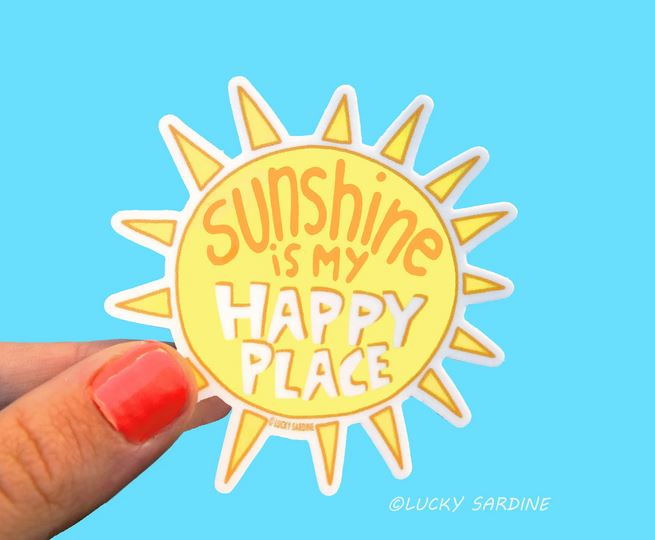 A hand with coral-colored nail polish holds the Sunshine Is My Happy Place sticker from Lucky Sardine. The waterproof vinyl sticker, shaped like a yellow sun with pointed rays, features the text "sunshine is my HAPPY PLACE" in a cheerful, playful font. The background is a solid bright blue color, and "@LUCKY SARDINE" appears in small credit text.