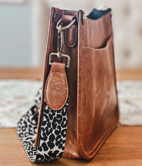 The Sydney Crossbody - Brown by zzzLivFoxy, with a detachable strap and featuring a black and white leopard print design, hangs on a wooden table. This vegan leather bag showcases sleek edges and a visible side pocket, set against a softly blurred background.