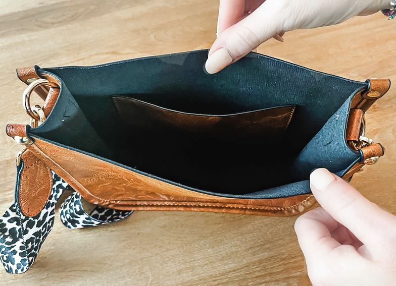 Hands opening the Sydney Crossbody - Brown by zzzLivFoxy, showcasing its vegan leather exterior and sleek black interior. This stylish bag features a leopard print strap and a small inner pocket, seamlessly combining style and practicality. It's beautifully displayed on a wooden surface.