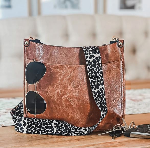 The Sydney Crossbody - Brown by zzzLivFoxy, featuring a leopard print strap, sits on a wooden table. Sunglasses are tucked into its pocket, and car keys rest beside it. In the background, a cushioned bench and framed pictures adorn the wall.