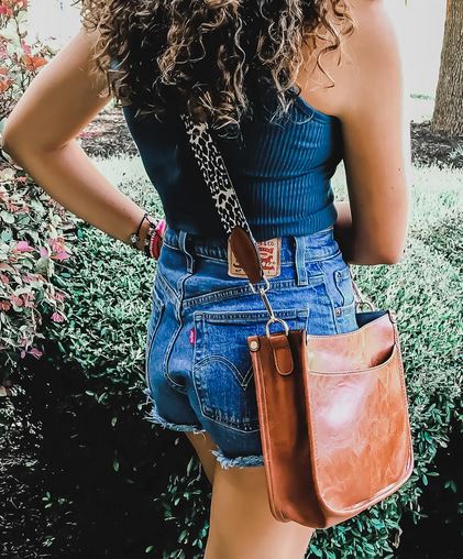 A person with curly hair stands outdoors, wearing a black tank top and denim shorts. They carry the stylish Sydney Crossbody in brown from zzzLivFoxy, featuring a chic vegan leather design and leopard-print strap. Lush green bushes and a tree trunk are visible in the background.