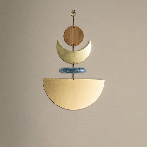 The Moonrise Labradorite Wall Hanging by Scout Jewelry is a minimalist piece of sculptural wall art featuring a wooden circle at the top, followed by a crescent moon shape. It is elegantly highlighted with a semi-precious stone-like blue bar in the center and concluded with a large natural brass half-circle at the bottom, all beautifully linked by a chain against a neutral wall.