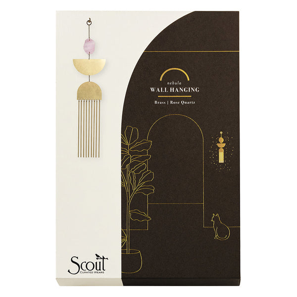Product packaging for a Scout Jewelry Nebula Rose Quartz Wall Hanging features a gold illustration of a plant, arch, candle, and cat. The sculptural wall art with brass and rose quartz details is elegantly displayed on the left.