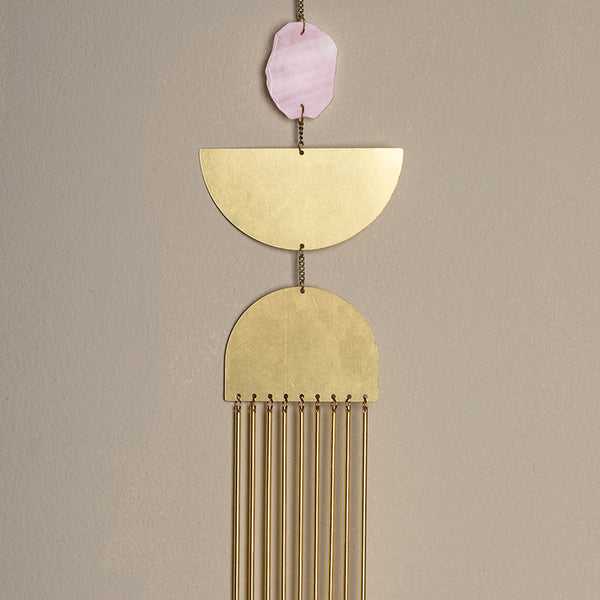 The Nebula Rose Quartz Wall Hanging by Scout Jewelry features a pink rose quartz accent, gold half-circle and semicircle shapes, and vertical brass rods suspended by chains, creating sculptural wall art against a beige backdrop.